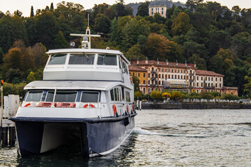Naklejka premium Modern catamaran ferry departs Lake Como, past historic grand buildings and lush autumn trees on the tranquil Italian shore. Picturesque landscape, classic charm, calm waters.