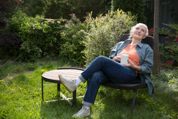 Fototapeta premium portrait of a cheerful elderly woman with a cup of tea in the garden about her home on a summer. concept without stress. mental health. Enjoying the little things. Mindfulness and feel-good moments.