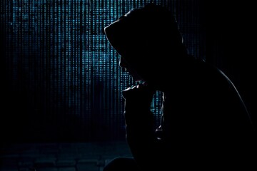 Mysterious hacker in hooded silhouette working in dark room, highlighting cybersecurity, data protection, and internet privacy themes.