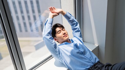 Young Man Relaxing in Bright Room by Window on Sunny Day with Hands Raised and Eyes Closed