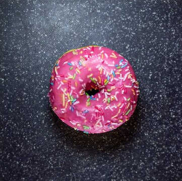A single pink glazed donut with colorful sprinkles on a dark textured surface
