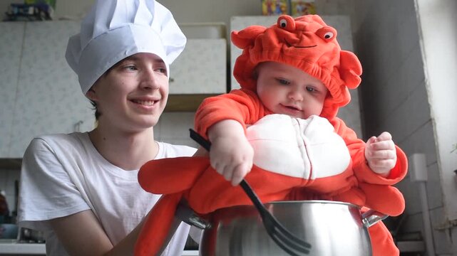  Children in carnival costumes in the kitchen. A teenage boy in a chef's hat sits next to his baby brother in a red crab or lobster costume sitting in a saucepan.