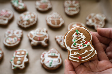 Caucasian hand holding homemade gingerbread cookie in the form of snow globe, Christmas gingerbread cookies of different shapes on baking paper on background, holiday food
