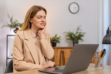 Middle-aged businesswoman at home office works on laptop feels tooth pain winces holds cheek tries...