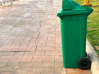 A tall, green trash can with black wheels. It's standing on a stone-paved walkway next to a grassy area. The walkway has some cracks in it and appears to be in a park or public space, with a concrete.