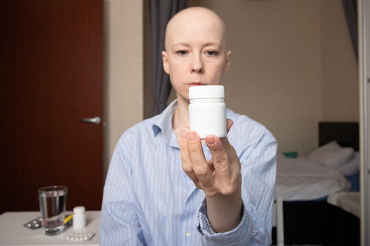 Bald woman in a striped shirt holding a white pill bottle with medicines and a glass of water on the table, concept of cancer treatment, chemotherapy therapy, patient taking drugs at home