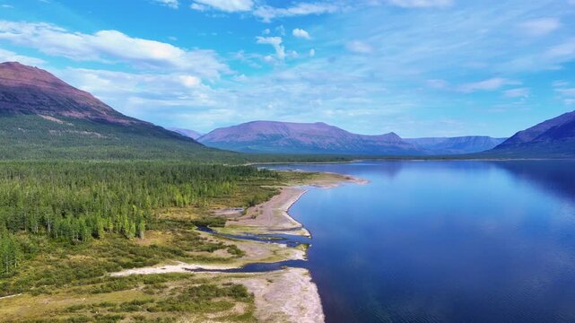Aerial view of Lake Lama coastline with green taiga and purple mountains of the Putorana Plateau in summer daylight. Clear water, long sandbars and scale create a rare travel destination in Siberia