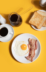 Fried egg with roasted bacon, black coffee, toasts on yellow background. Retro American breakfast. Hard shadow.