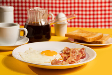 Fried egg with roasted bacon, black coffee, toasts on yellow background. Retro American breakfast. Hard shadow.