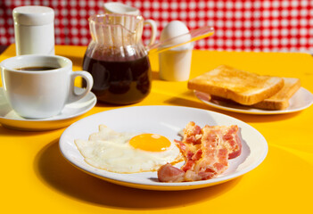 Fried egg with roasted bacon, black coffee, toasts on yellow background. Retro American breakfast. Hard shadow.