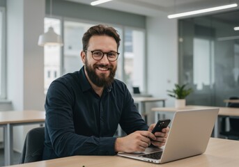 Fototapeta premium Smiling bearded man with glasses holding a smartphone while sitting at a desk with a laptop in a modern office environment