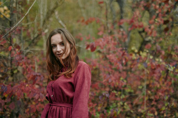 A candid portrait of a young woman in a burgundy dress among autumn foliage, offering a genuine, authentic expression and natural pose captured with warm earthy tones and soft focus.