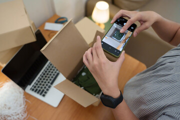 Asian woman taking photo of packed clothes inside cardboard box with smartphone before shipping for online shop. Small business concept.