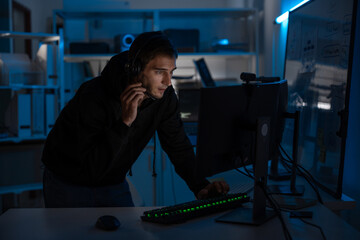 Male programmer and developer focused on computer screen at night, coding digital systems and ensuring cyber security. Freelancer with headset working overtime under blue light in office.