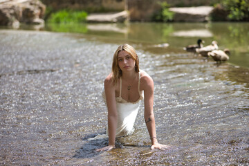 Young, beautiful, blonde woman in a white dress, crawling on all fours over a small waterfall in a lake with ducks in an outdoor park. Concept of beauty, youth, millennial.