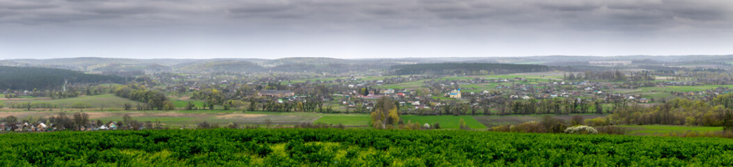 Fototapeta premium A panoramic view of Mliyiv village, Cherkasy region, Ukraine, in the Vilshanka River valley, during spring late April. The photo shows houses and buildings surrounded by green fields and rolling hills