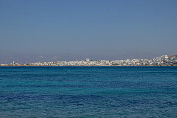 View of a whitewashed coastal town on a Greek island with turquoise sea in the foreground and mountains in the background.
