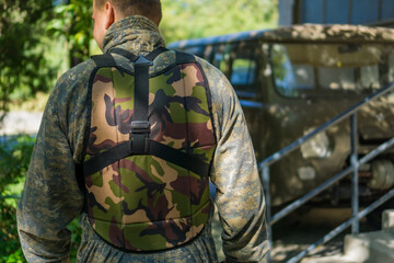 Back view of a man wearing a camouflage vest or chest protector, the man is standing in an outdoor military or airsoft game area, with a vehicle in the background, concept of adventure, tactics, hobby