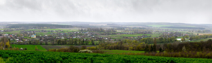 Fototapeta premium A panoramic view of Mliyiv village, Cherkasy region, Ukraine, in the Vilshanka River valley, during spring late April. The photo shows houses and buildings surrounded by green fields and rolling hills