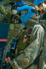 Paintball player wearing camouflage uniform and blue protective mask, holding a paintball marker, preparing for an outdoor match with teammates.