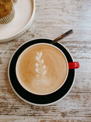 Overhead view of a latte with leaf art, on a black saucer, with a muffin. A crafted cappuccino next to a muffin, on a rustic wooden table. The overhead shot captures the details. Morning drink.