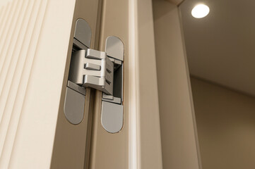 A close-up of a modern door hinge, showcasing its sleek, concealed design within a light-colored wooden door. A bright ceiling light is visible in the background