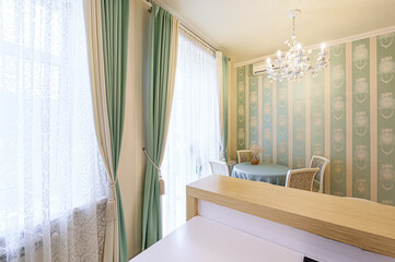 A bright, elegant room features mint and cream curtains framing a window. A round table with chairs sits against striped wallpaper, illuminated by a chandelier