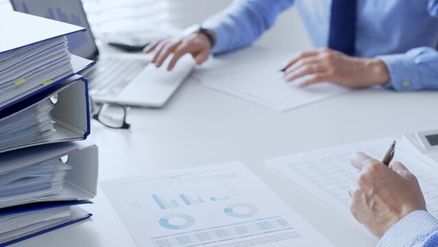 Team of accountants or auditors are analyzing financial reports and making notes while sitting at a white table, close up of a stack of blue folders full of paperwork. Audit and taxes theme in