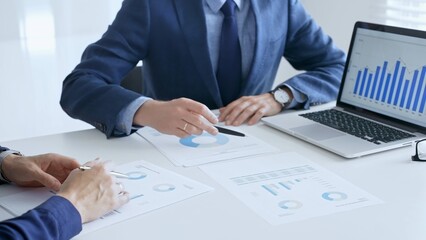 Business colleagues or partners are discussing financial data on a laptop and printed documents while analyzing charts and market trends at office table. Businesspeople and finance concept