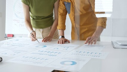 Two business professionals are examining financial data and reports, pointing at charts and graphs displayed on a table during a collaborative office meeting