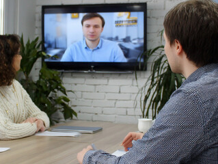 Colleagues participating in virtual meeting, watching male speaker on screen during video conference in modern office with white brick wall and indoor plants
