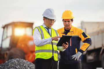 Professionals in Safety Gear Review Project Plans on Tablet at Construction Site with Heavy Machinery and Gravel Background