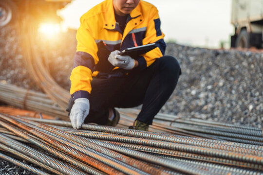 Construction worker inspecting steel rods at a construction site, wearing safety gear and holding a clipboard in the daylight with a blurred background