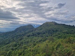 Fototapeta premium Mountain Landscape under a Cloudy Sky: Majestic mountains covered in lush greenery stand tall against a dramatic sky filled with clouds, embodying nature's grandeur and serene atmosphere.