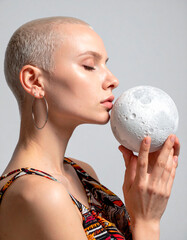 Naklejka premium A serene conceptual portrait of a person with shaved hair and light skin, eyes closed, gently holding a realistic moon model close to their face. Portrait with emotional depth and celestial symbolism.