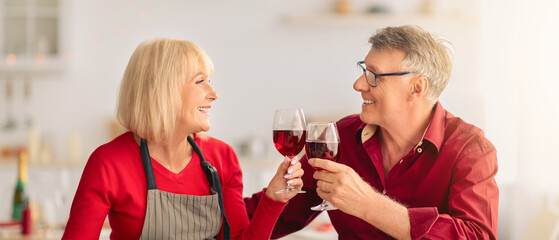 Loving senior couple having romantic times, drinking red wine while cooking festive Thanksgiving or Christmas dinner at kitchen. Elderly man and his wife enjoying alcoholic drink at home