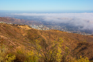 A wide-angle, high-angle view captures the scenic Pacific Palisades coastline from a dry hillside trail, with a low-lying marine layer of fog rolling over the Pacific Ocean below a clear blue sky in S