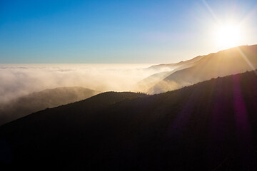 A wide-angle, high-angle view captures the scenic Pacific Palisades coastline from a dry hillside trail, with a low-lying marine layer of fog rolling over the Pacific Ocean below a clear blue sky in S