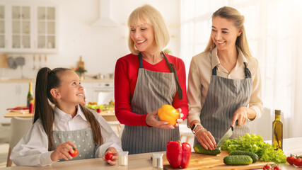 Happy multigeneration family cooking together, cutting vegetables for festive dinner at kitchen. Cheerful little girl with her mother and granny preparing healthy meal at home