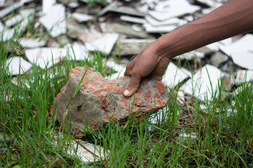 A hand picks up a broken brick from the grass near rubble