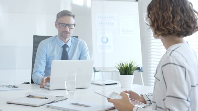 Business colleagues working together at sleek office desk, laptop and tablet open, paperwork spread, conveying professional teamwork and productivity in contemporary workspace - Powered by Adobe