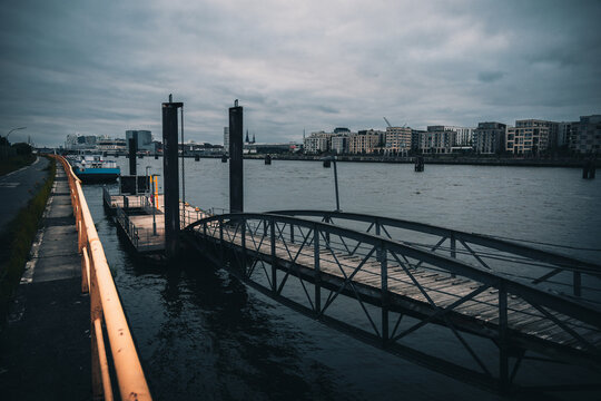Dock walkway leading to floating platform on urban river with overcast sky and waterfront skyline