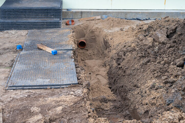 Construction Trench with Exposed Orange PVC Drain Pipe and Protective Mats on Sandy Worksite.
