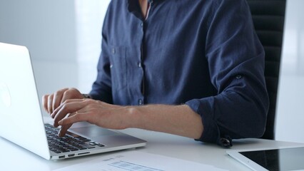 Businessman wearing a blue shirt is typing on a laptop with financial reports and a tablet on his...