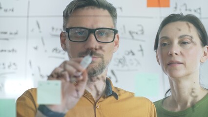 Professional team members brainstorming strategy, writing notes on transparent whiteboard with colorful sticky papers during collaborative meeting. Business people concept