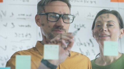 Professional team members brainstorming strategy, writing notes on transparent whiteboard with colorful sticky papers during collaborative meeting. Business people concept