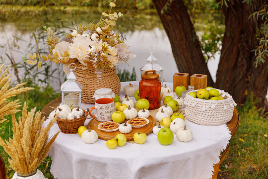 Rustic autumn table setting is arranged outdoors, next to a calm river. The round table is covered with a white cloth and is laden with seasonal harvest decor. - Powered by Adobe