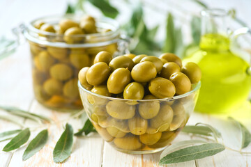 pickled green olives in a transparent glass bowl on a white wooden table, selective focus.