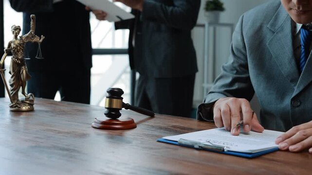 Lawyer in suit and tie sitting at desk with gavel and statue of Lady Justice, reviewing and signing a contract while another person stands in background - Powered by Adobe