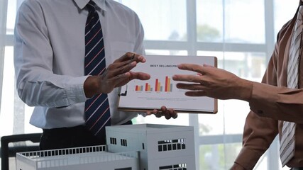 Professional architect presenting best selling product statistics on clipboard while standing near building scale model, sharing insights with colleagues in bright, modern office workspace - Powered by Adobe